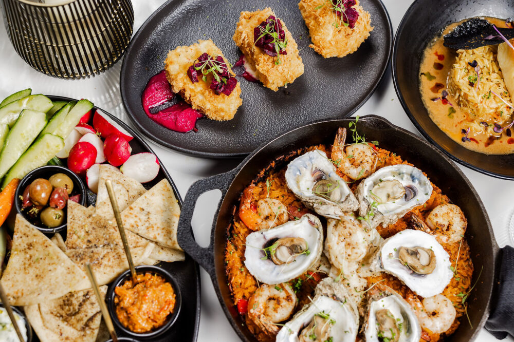 Four plates of food including oysters on the half-shell, pita and crudité, and two other dishes.